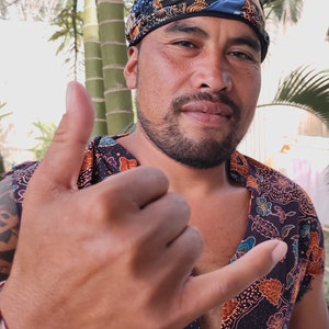 May include: A man with a dark beard and a colorful patterned bandana on his head is making the shaka sign with his hand. He is wearing a dark blue shirt with a colorful floral pattern.