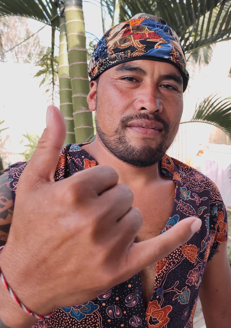 May include: A man with a dark beard and a colorful patterned bandana on his head is making the shaka sign with his hand. He is wearing a dark blue shirt with a colorful floral pattern.