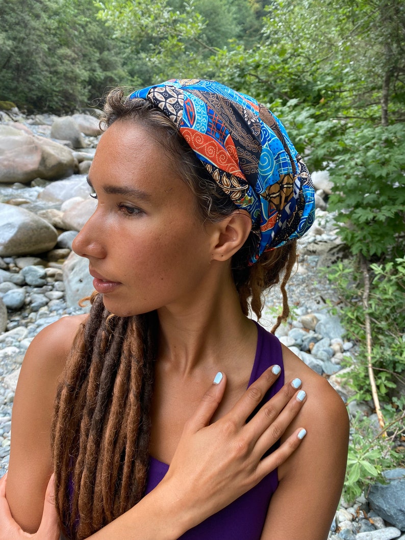 May include: A woman with long brown dreadlocks is wearing a blue, orange, and brown patterned headband. The headband is tied in a knot at the top of her head. She is wearing a purple tank top and has white nail polish on her nails.