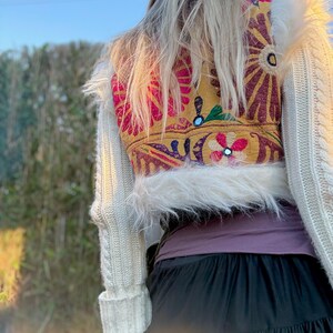 K&ouml;nnte beinhalten: Ein cremefarbener Zopfstrickpullover mit Kunstfellbesatz. Auf der R&uuml;ckseite des Pullovers befindet sich ein farbenfrohes, besticktes Panel mit Blumenmustern in Rosa, Gelb und Rot. Am unteren Bildrand ist ein schwarzer Rock zu sehen.