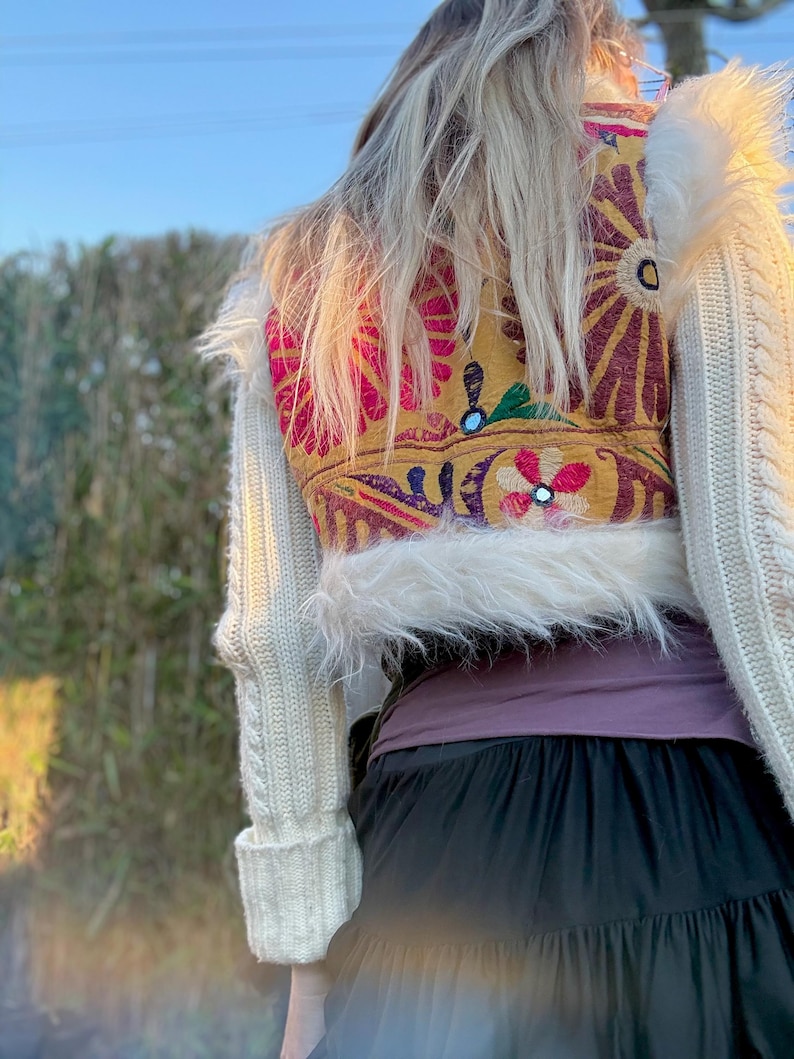 K&ouml;nnte beinhalten: Ein cremefarbener Zopfstrickpullover mit Kunstfellbesatz. Auf der R&uuml;ckseite des Pullovers befindet sich ein farbenfrohes, besticktes Panel mit Blumenmustern in Rosa, Gelb und Rot. Am unteren Bildrand ist ein schwarzer Rock zu sehen.