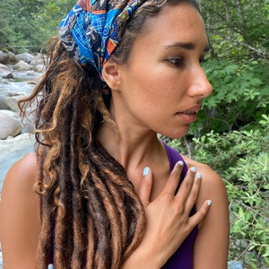 May include: A person with long brown dreadlocks wearing a blue and orange patterned headband. The person is wearing a purple tank top and has their hand on their chest.