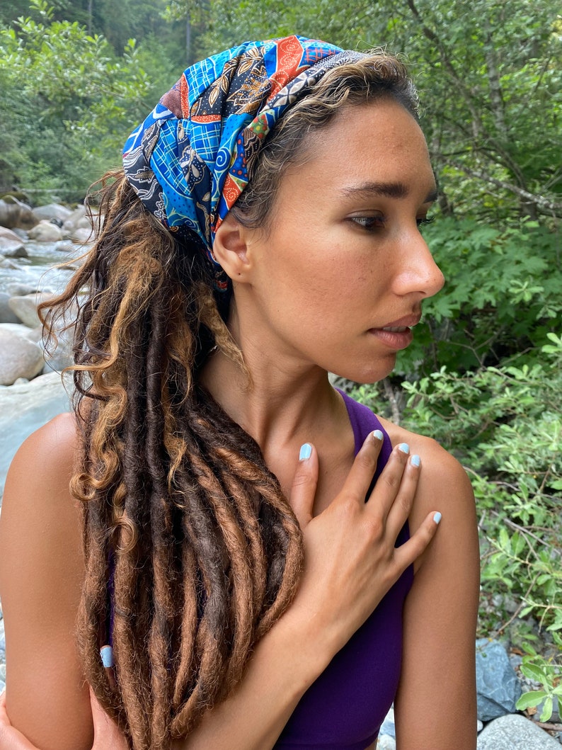 May include: A person with long brown dreadlocks wearing a blue and orange patterned headband. The person is wearing a purple tank top and has their hand on their chest.