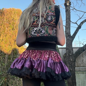 May include: A sleeveless vest featuring black fabric with colorful embroidered patterns and small circular mirrors, paired with a layered skirt consisting of a deep red patterned fabric, a shiny purple satin tier, and a black ruffled hem.