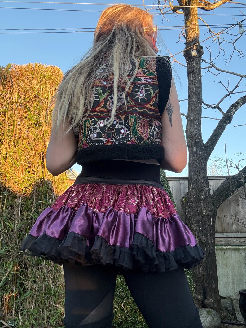 May include: A sleeveless vest featuring black fabric with colorful embroidered patterns and small circular mirrors, paired with a layered skirt consisting of a deep red patterned fabric, a shiny purple satin tier, and a black ruffled hem.