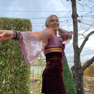May include: A woman wearing a purple and orange sheer top with a brown velvet skirt. The top has a gold and purple trim and a tie at the waist. The woman is standing in front of a green bush and a tree.