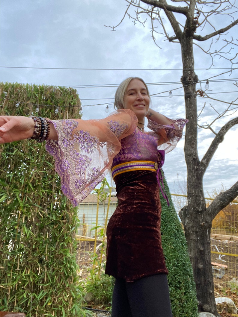 May include: A woman wearing a purple and orange sheer top with a brown velvet skirt. The top has a gold and purple trim and a tie at the waist. The woman is standing in front of a green bush and a tree.