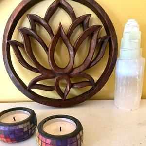 May include: A wooden wall hanging with a carved lotus flower design. The hanging is brown and has a circular shape. Two mosaic candles are in the foreground.