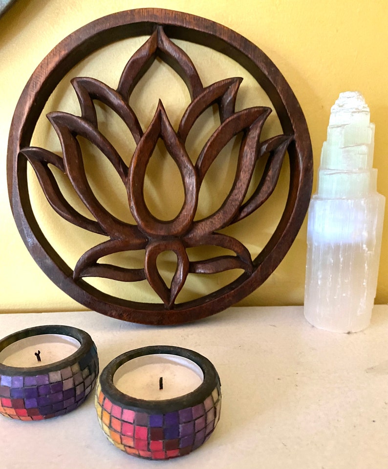 May include: A wooden wall hanging with a carved lotus flower design. The hanging is brown and has a circular shape. Two mosaic candles are in the foreground.