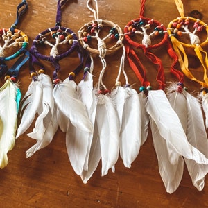 May include: A set of six colourful dreamcatchers with white feathers. Each dreamcatcher features a beaded tree of life design and is strung with colourful beads.