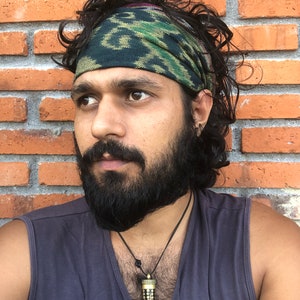 May include: A man with a dark beard and long, curly hair is wearing a green, black, and gold patterned bandana around his head. He is wearing a gray tank top and a necklace with a black and gold pendant. He is standing in front of a brick wall.