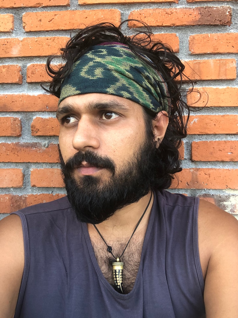 May include: A man with a dark beard and long, curly hair is wearing a green, black, and gold patterned bandana around his head. He is wearing a gray tank top and a necklace with a black and gold pendant. He is standing in front of a brick wall.