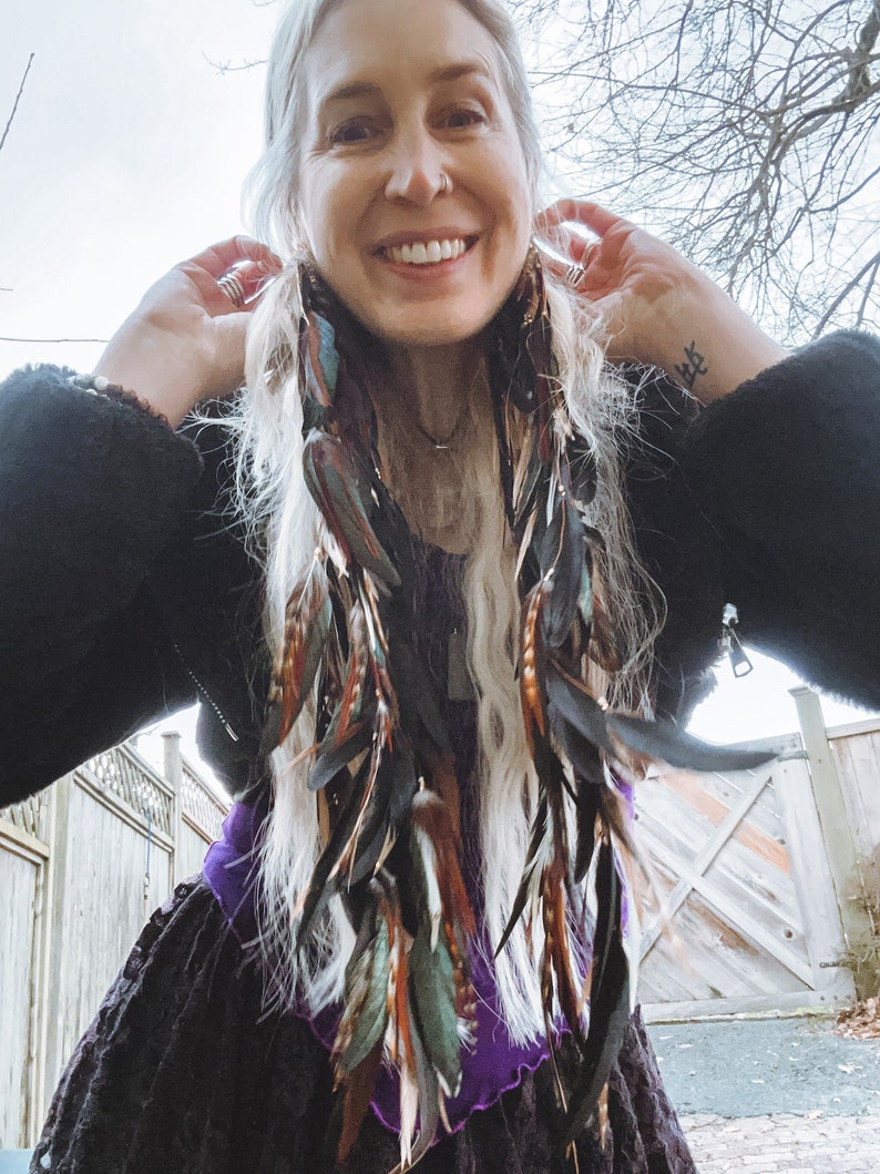 May include: A woman with long, white hair wearing a black jacket and a purple and black dress with long feather earrings. The earrings are made of black, brown, and teal feathers.