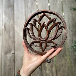 May include: A round wooden wall hanging with a carved lotus flower design. The wood is a dark brown color and the carving is intricate.