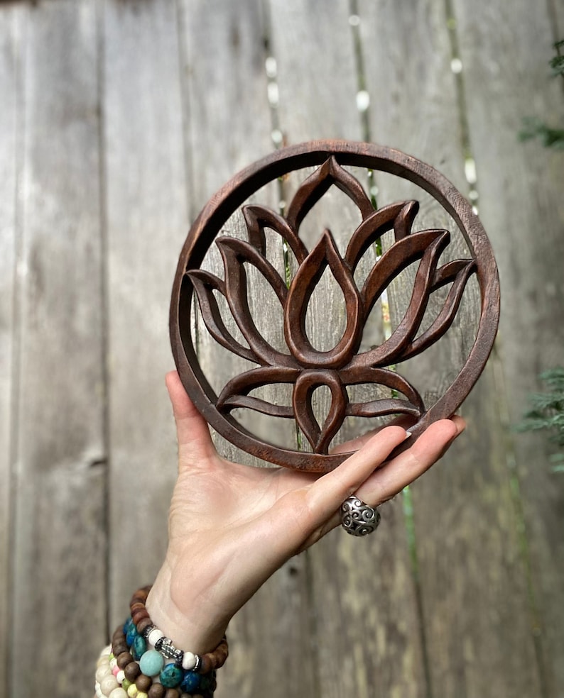 May include: A round wooden wall hanging with a carved lotus flower design. The wood is a dark brown color and the carving is intricate.