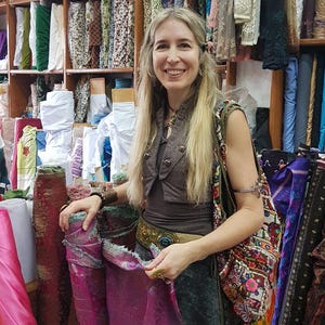 May include: A person with long blonde hair wearing a brown vest, a dark brown top, and a dark skirt, holding a piece of magenta fabric. The person is accessorized with a wide belt, a large patterned shoulder bag, and metal arm cuffs.