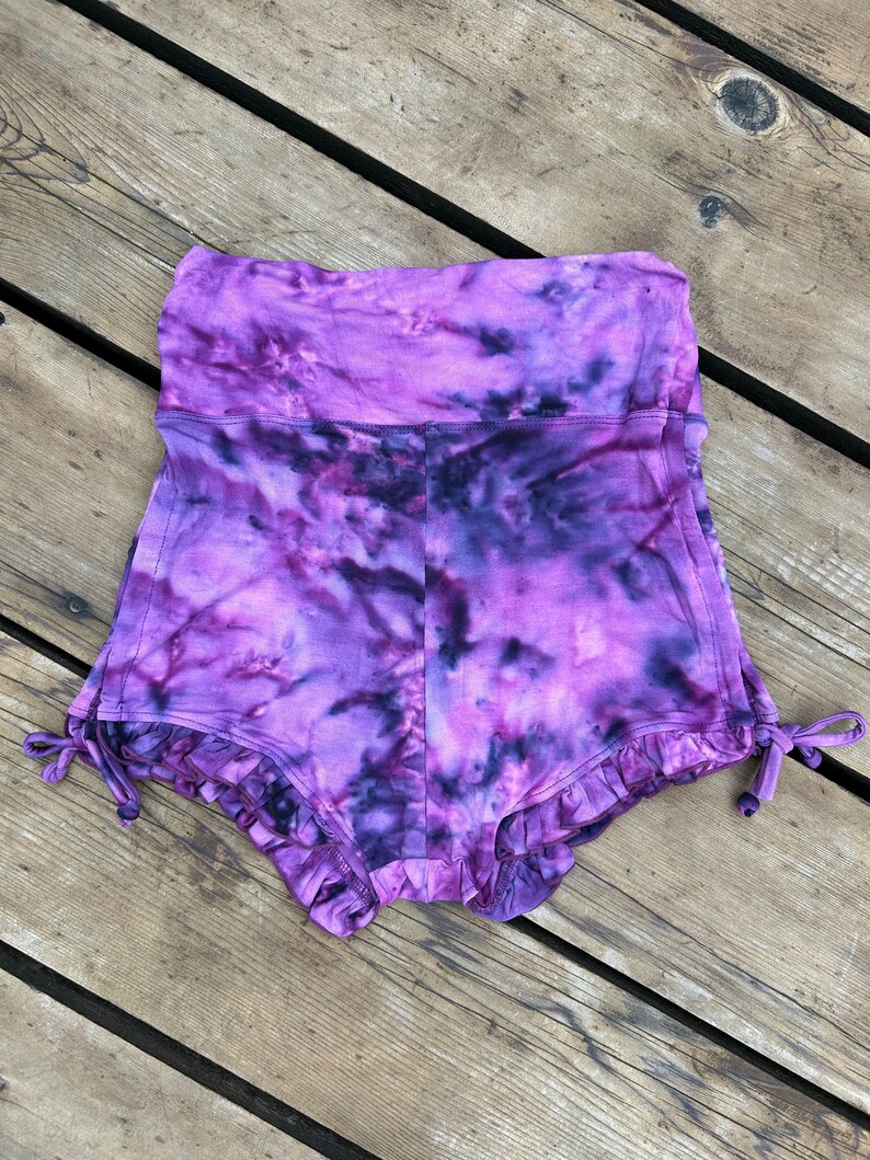 Puede incluir: Shorts de color morado tie-dye con un dobladillo con volantes y cordones de ajuste en los lados.