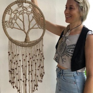 May include: A macrame wall hanging in the shape of a tree with brown wooden beads. The wall hanging is made of natural fibre rope and has a circular frame.