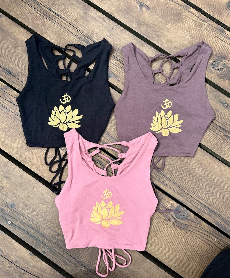 May include: Three tank tops with a lotus flower and Om symbol design. The tank tops are black, brown, and pink. The lotus flower and Om symbol are gold.