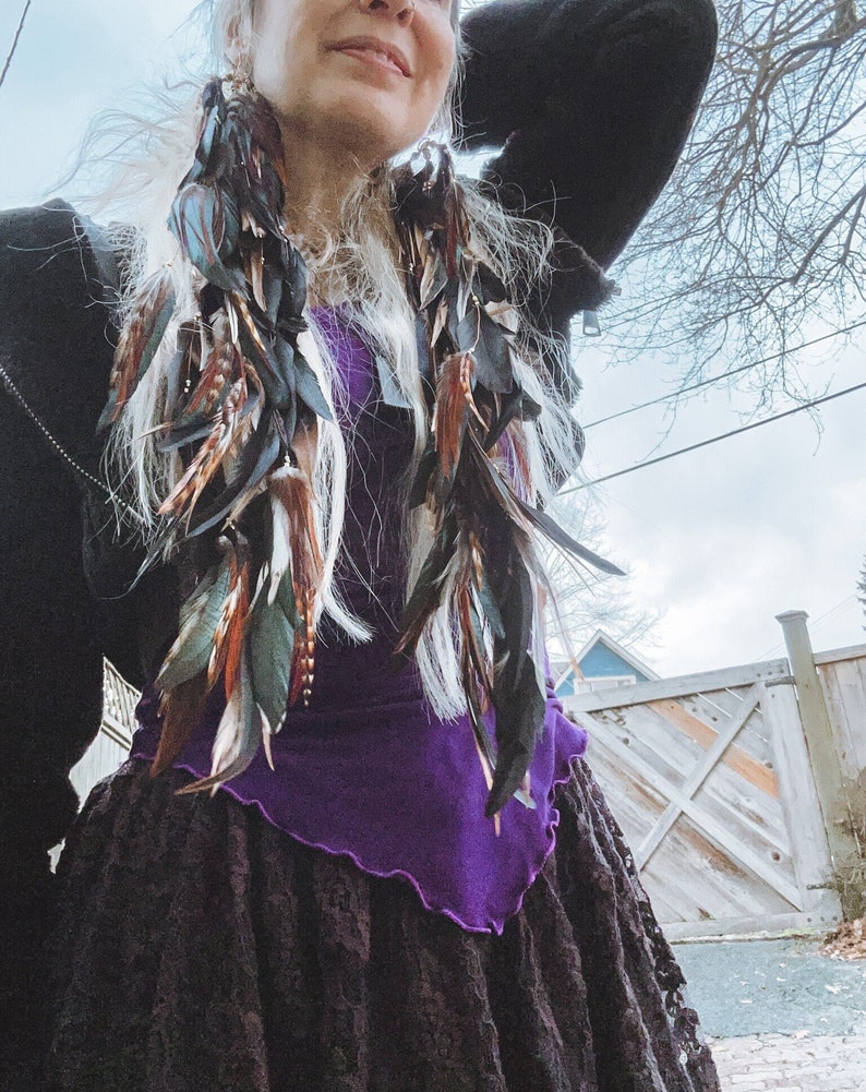 May include: A woman with long white hair wearing a black jacket, a purple top, and a black lace skirt. She is wearing long feather earrings with brown, green, and black feathers.