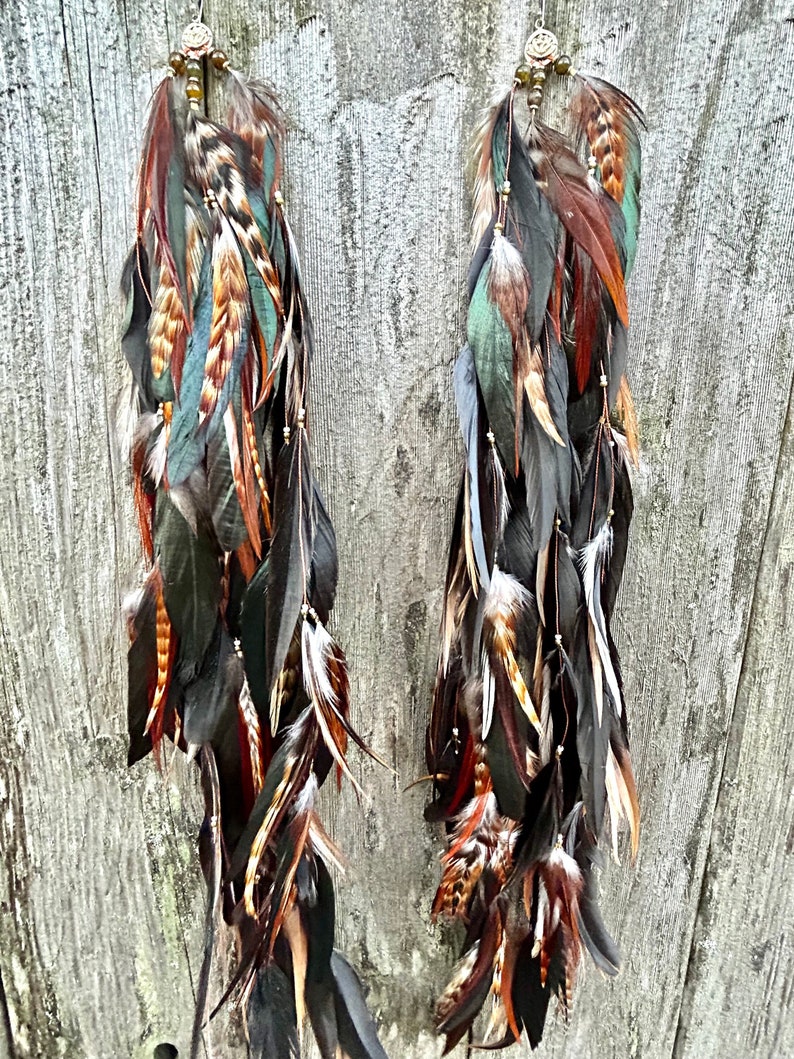 May include: A pair of long feather earrings with brown, black, green, and white feathers. The earrings have a beaded tassel and a metal charm at the top.