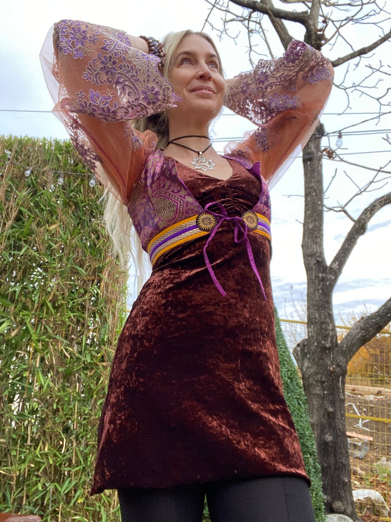 May include: A brown velvet mini dress with a purple and gold belt and a purple lace overlay. The dress has long sleeves with a purple and gold lace pattern. The model is wearing a silver necklace with a star pendant.
