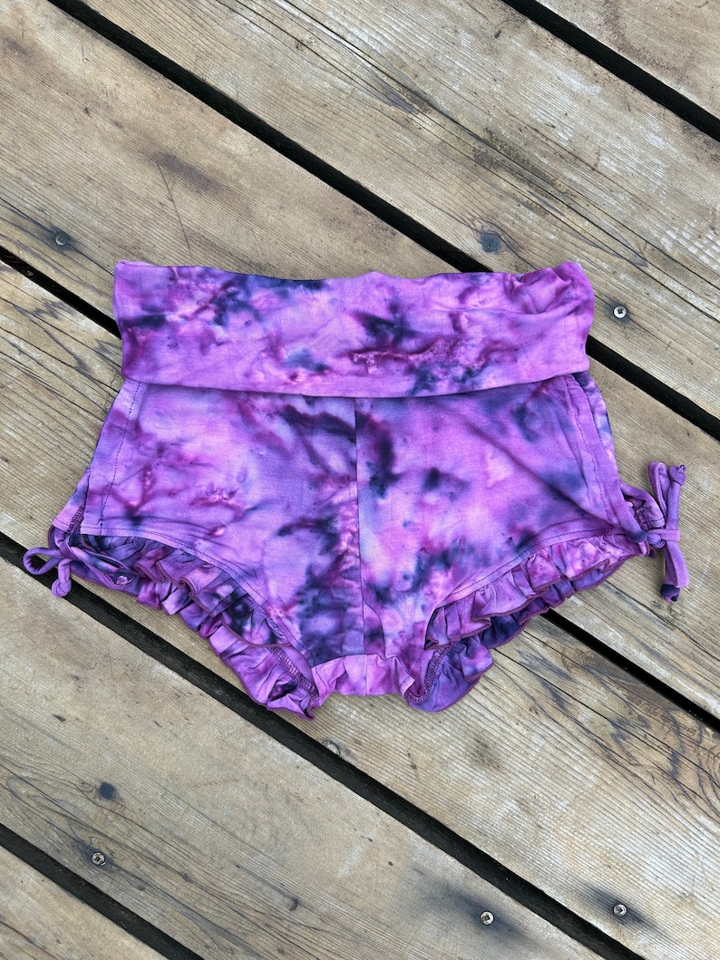 Puede incluir: Shorts de color morado con estampado tie-dye, dobladillo con volantes y un lazo en el lateral.