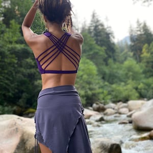 May include: A woman wearing a purple sports bra with a criss-cross back design and a gray wrap skirt. She is standing in a forest setting with a river in the background.