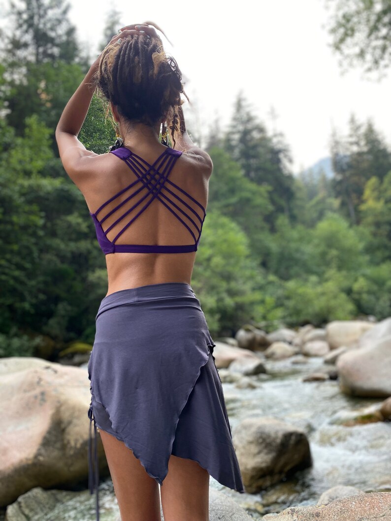 May include: A woman wearing a purple sports bra with a criss-cross back design and a gray wrap skirt. She is standing in a forest setting with a river in the background.