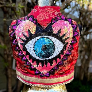 Puede incluir: Chaleco rojo con un diseño de ojo en forma de corazón. El ojo está hecho de lentejuelas azules, blancas y negras, rodeado de lentejuelas rosas y moradas. El chaleco tiene un ribete dorado y un estampado floral.