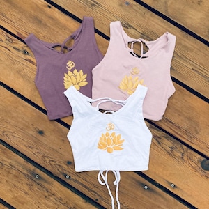 May include: Three crop tops with a lotus flower and Om symbol design. The tops are in white, purple, and pink.