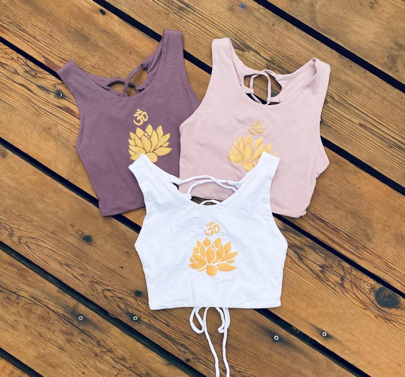 May include: Three crop tops with a lotus flower and Om symbol design. The tops are in white, purple, and pink.