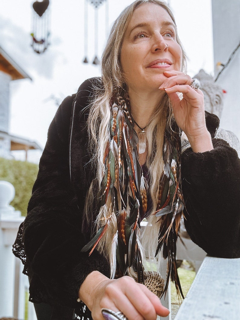 May include: A woman with long blonde hair wearing a black jacket with a zipper and a feather hair extension. The hair extension is made of brown, white, and green feathers. She is wearing a necklace with a large crystal pendant.