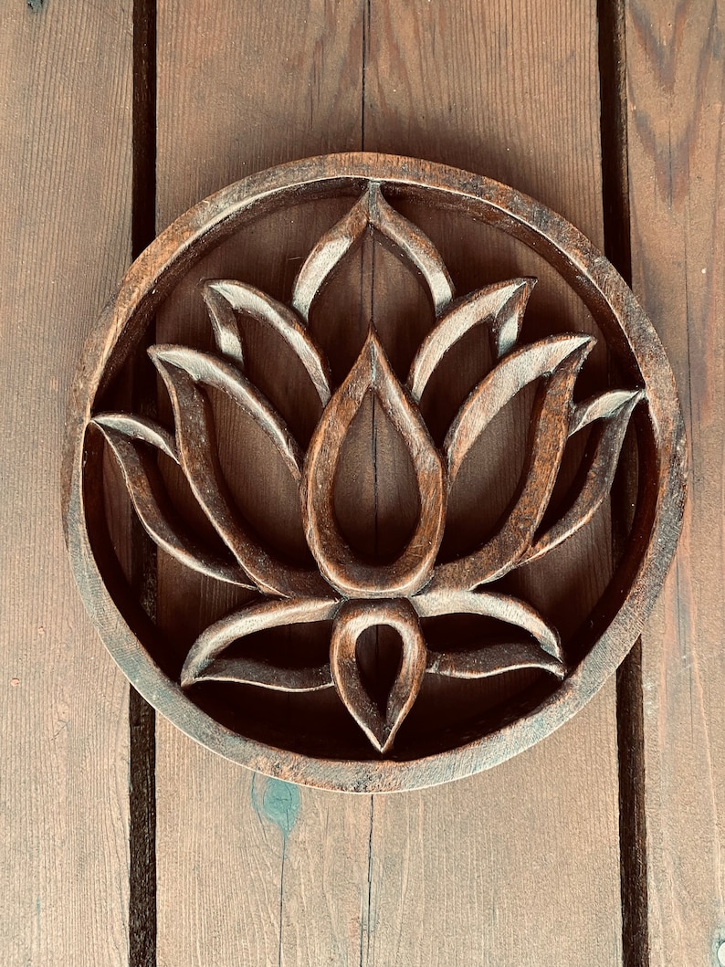May include: A round wooden wall decor with a carved lotus flower design. The wood is a dark brown color and the carving is intricate.