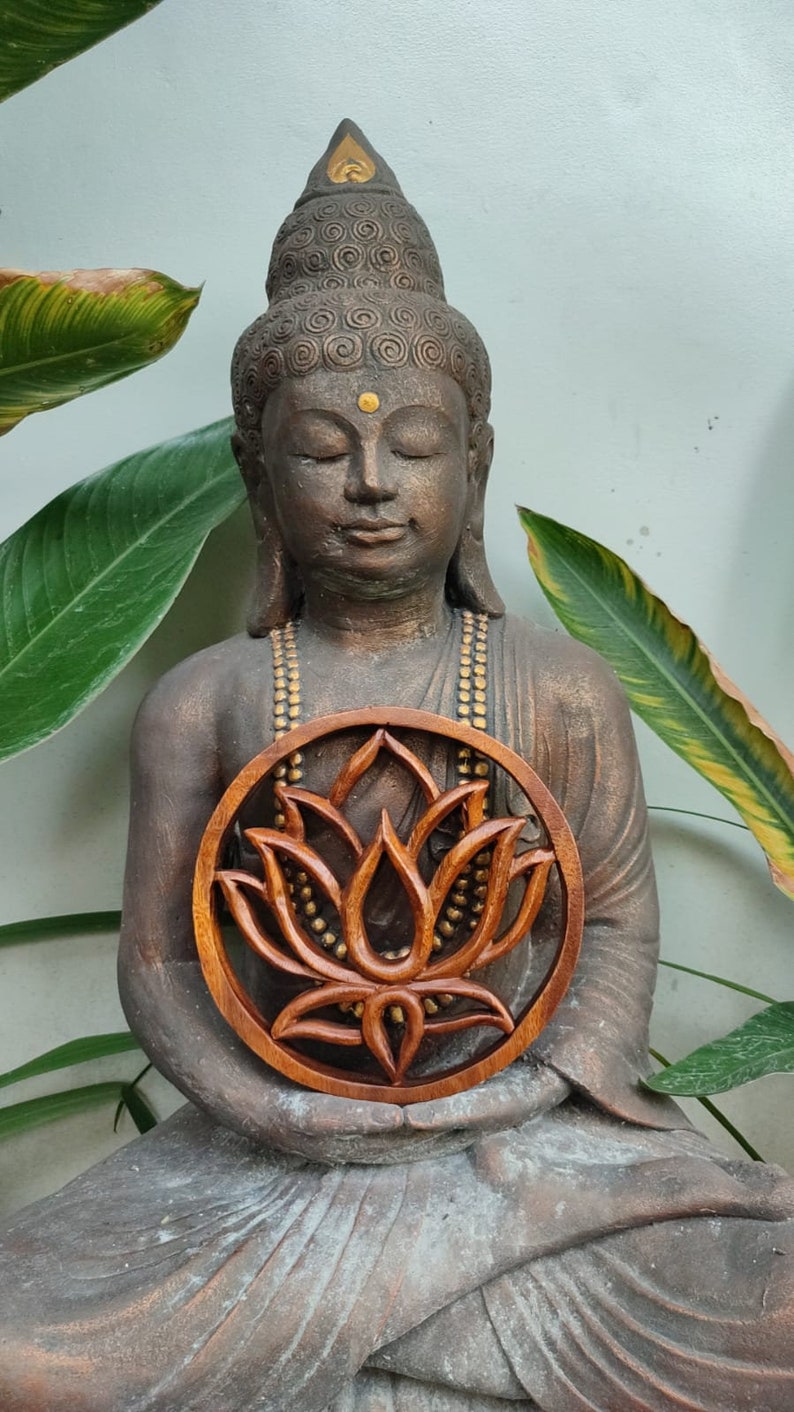 May include: A brown wooden statue of a meditating Buddha holding a round wooden carving of a lotus flower with gold accents.