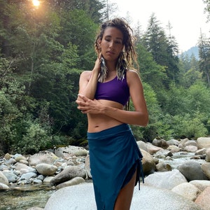May include: A woman wearing a purple sports bra and a teal blue wrap skirt with a long, braided hairstyle. She is standing in a forest setting with rocks and a stream in the background.