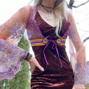 May include: A brown velvet dress with a purple and gold patterned overlay and a purple ribbon tie. The dress has long sleeves with lace detailing and a silver necklace with a star-shaped pendant.