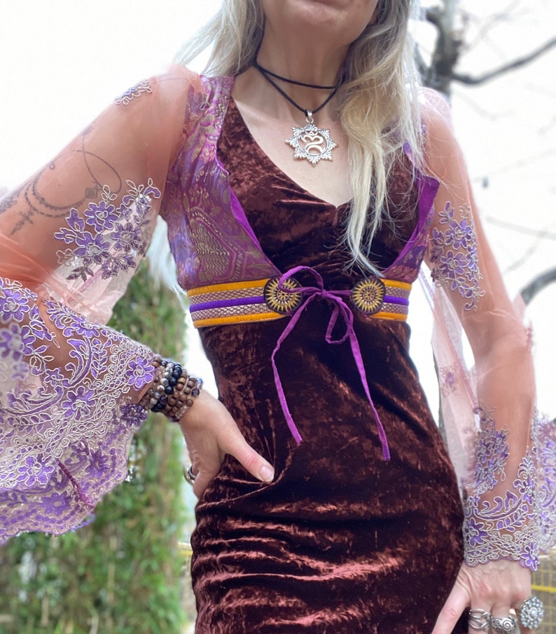 May include: A brown velvet dress with a purple and gold patterned overlay and a purple ribbon tie. The dress has long sleeves with lace detailing and a silver necklace with a star-shaped pendant.
