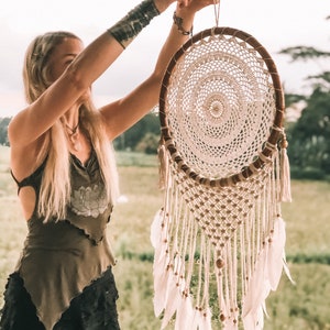 May include: A white dreamcatcher with brown wooden beads and white feathers hanging from a brown wooden hoop. The dreamcatcher is being held by a person.