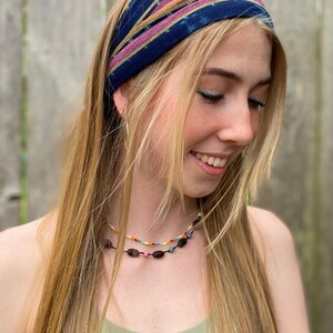 May include: A woman with long blonde hair is wearing a blue and pink patterned headband. She is also wearing a green tank top and a beaded necklace with dark brown oval beads.