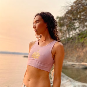 May include: A woman wearing a light purple crop top with a gold lotus flower design. The top is sleeveless and has a scoop neck. The woman is standing in front of a body of water and a rocky shoreline.