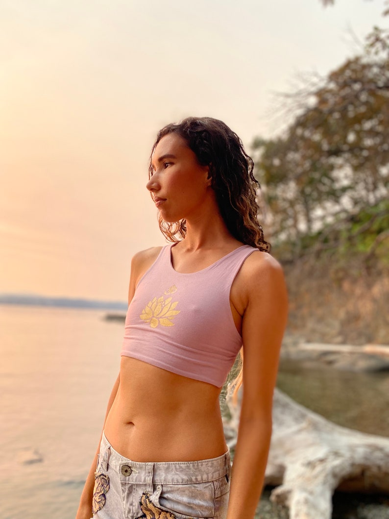 May include: A woman wearing a light purple crop top with a gold lotus flower design. The top is sleeveless and has a scoop neck. The woman is standing in front of a body of water and a rocky shoreline.