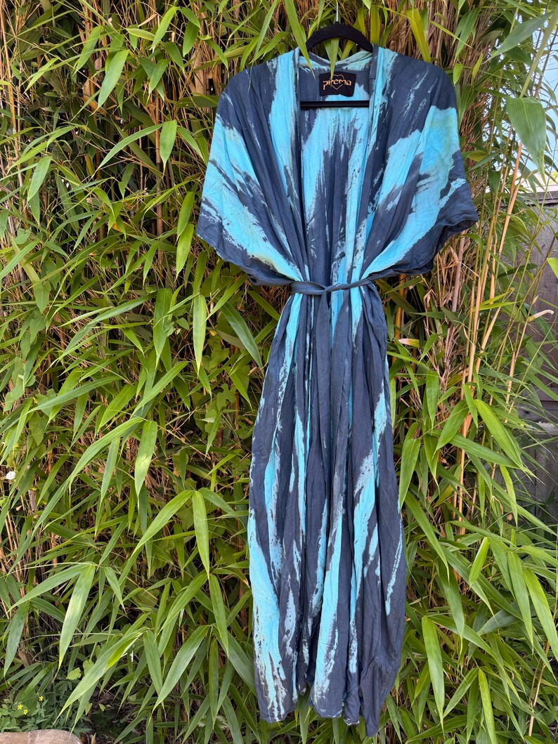 Peut inclure: Une robe longue ouverte sur le devant avec une fermeture &agrave; nouer &agrave; la taille et un motif tie-dye gris anthracite fonc&eacute; et bleu turquoise vif. Le v&ecirc;tement pr&eacute;sente des manches courtes et larges ainsi qu'une &eacute;tiquette noire au col avec le texte "Pirema" en dor&eacute;.