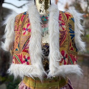 K&ouml;nnte beinhalten: Eine bestickte Weste mit wei&szlig;em Kunstfellbesatz. Die Weste hat einen goldenen Hintergrund und zeigt florale Muster in Pink, Blau und Rot. Die Weste ist Teil eines Outfits im Boh&egrave;me-Stil.