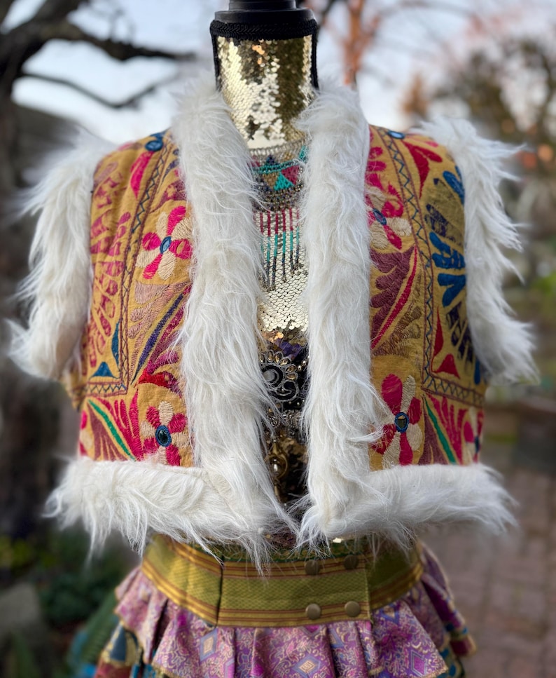 K&ouml;nnte beinhalten: Eine bestickte Weste mit wei&szlig;em Kunstfellbesatz. Die Weste hat einen goldenen Hintergrund und zeigt florale Muster in Pink, Blau und Rot. Die Weste ist Teil eines Outfits im Boh&egrave;me-Stil.