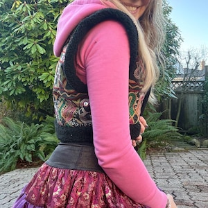May include: A pink hooded sweatshirt worn under a black sleeveless vest with colorful embroidered patterns and small mirrors. The outfit includes a dark waistband and a layered skirt featuring a maroon floral print and a purple satin ruffle hem.