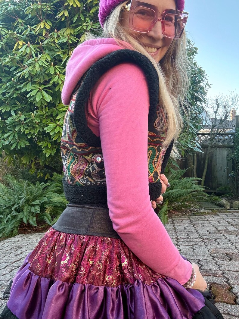 May include: A pink hooded sweatshirt worn under a black sleeveless vest with colorful embroidered patterns and small mirrors. The outfit includes a dark waistband and a layered skirt featuring a maroon floral print and a purple satin ruffle hem.