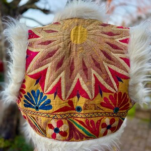 Colorful, embroidered vest with a sunburst design in gold, red, and pink. White faux fur trim and floral and geometric patterns.