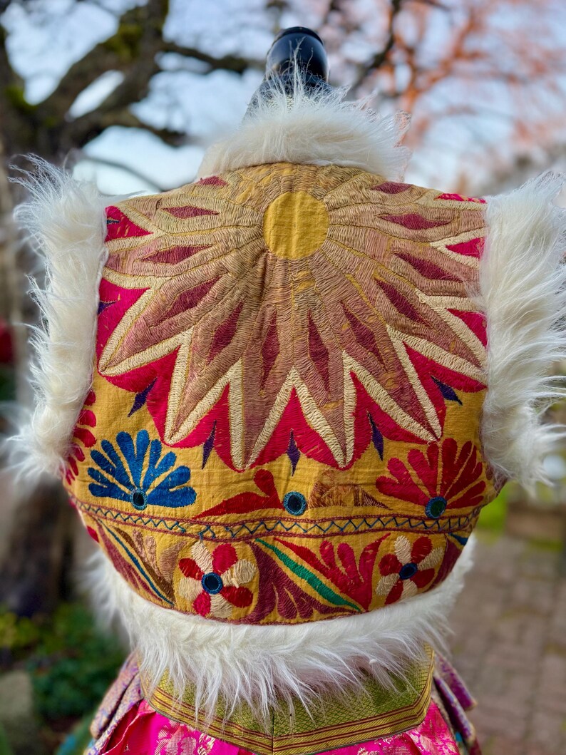 Colorful, embroidered vest with a sunburst design in gold, red, and pink. White faux fur trim and floral and geometric patterns.