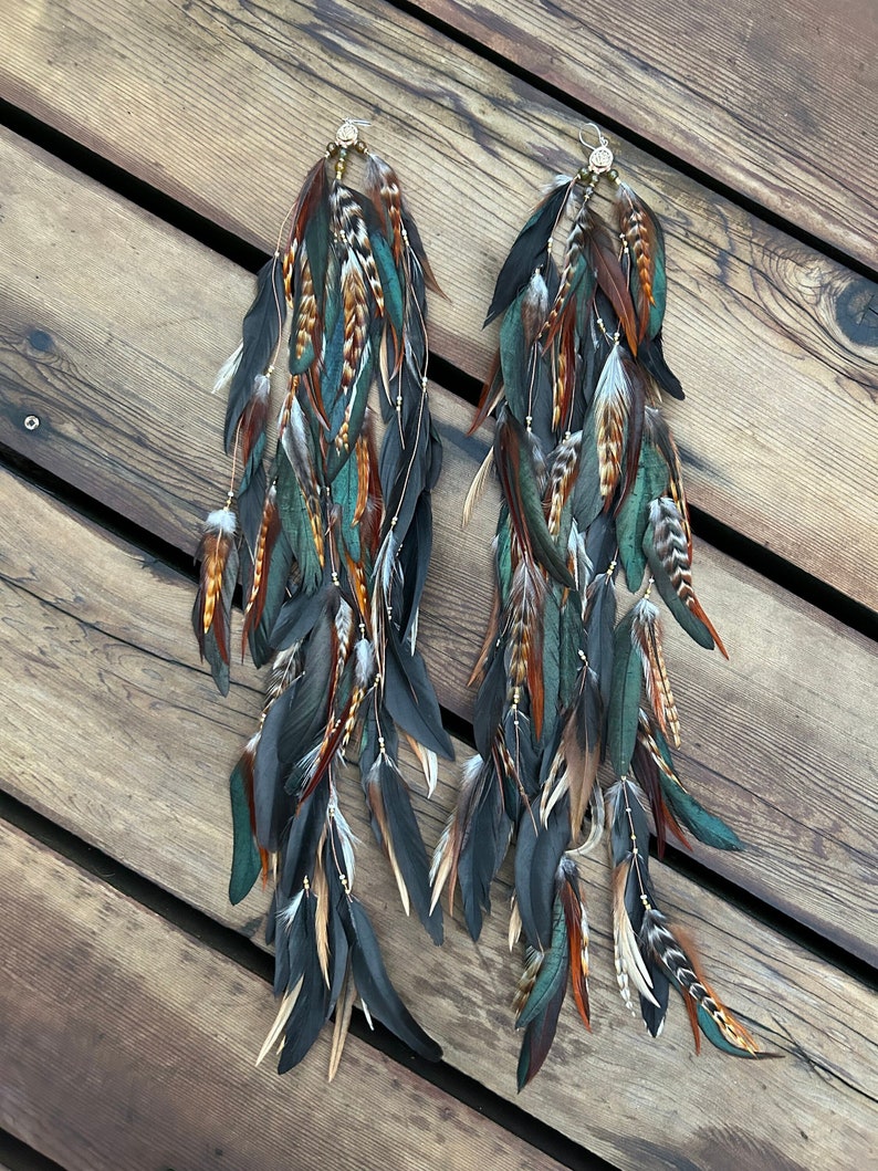 A pair of long, dangling earrings with feathers in shades of brown, teal, and black. The earrings are made with a variety of feathers some with a sheen.